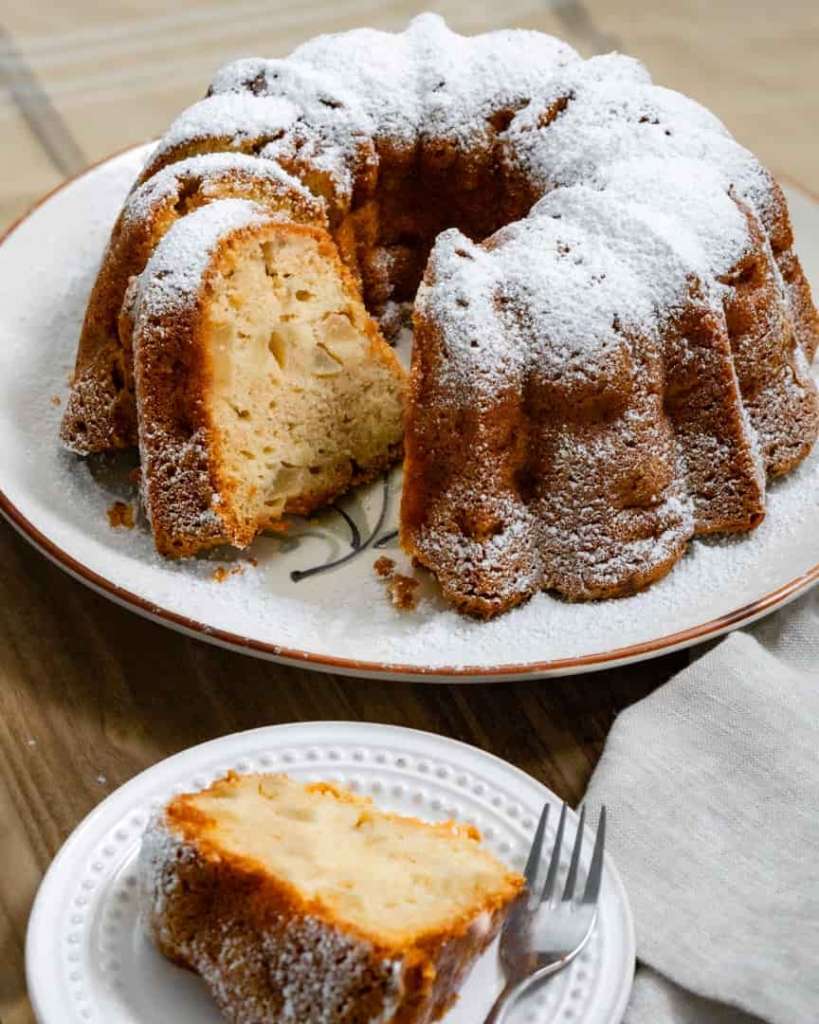 A slice of bundt cake on a plate with a fork. A bundt cake with the piece missing placed behind it.