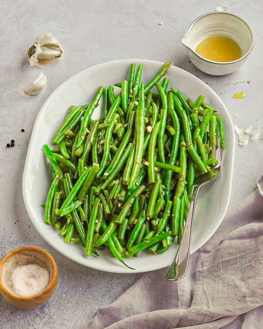 Green beans on a platter with a small pitcher of olive oil, garlic clove and small bowl of salt next to it.