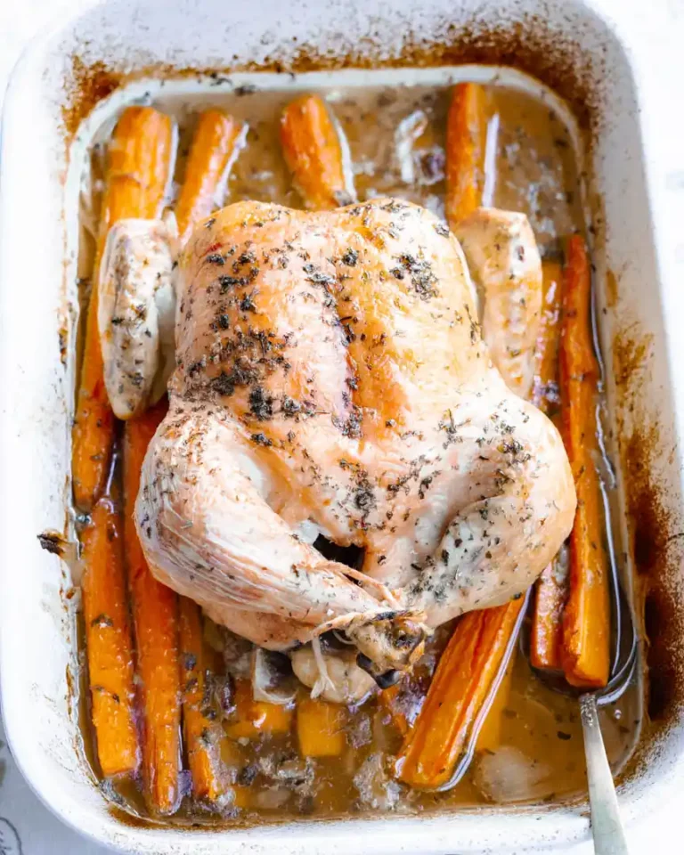 Roasted chicken over carrots in a baking pan. There is a spoon sitting in the pan to scoop up pan juices.