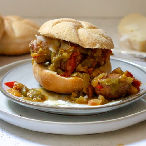 Italian Sausage & Peppers sandwich on a plate. There is some spilled over sausage bites and peppers next to the sandwich on the plate.