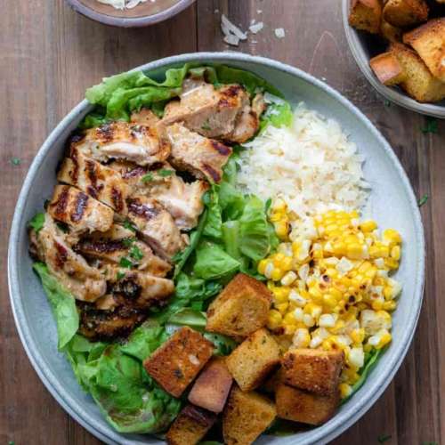A bowl with grilled chicken sliced on a bed of lettuce. And sectioned piles around the bowl with croutons, grilled corn, shredded cheese.