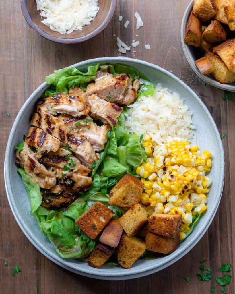 A bowl with grilled chicken sliced on a bed of lettuce. And sectioned piles around the bowl with croutons, grilled corn, shredded cheese.