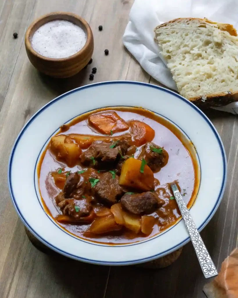 A bowl of beef stew with carrots and potatoes and sliced bread on the side