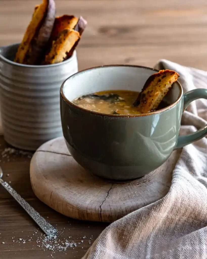 Escarole, bean & Italian sausage soup in a mug with a piece of garlic bread dipped into the soup