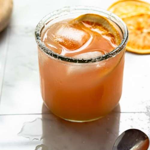 Paloma cocktail in a short glass with ice cubes and dried orange slice floating on top. With dried orange slices and a spoon next to the glass.