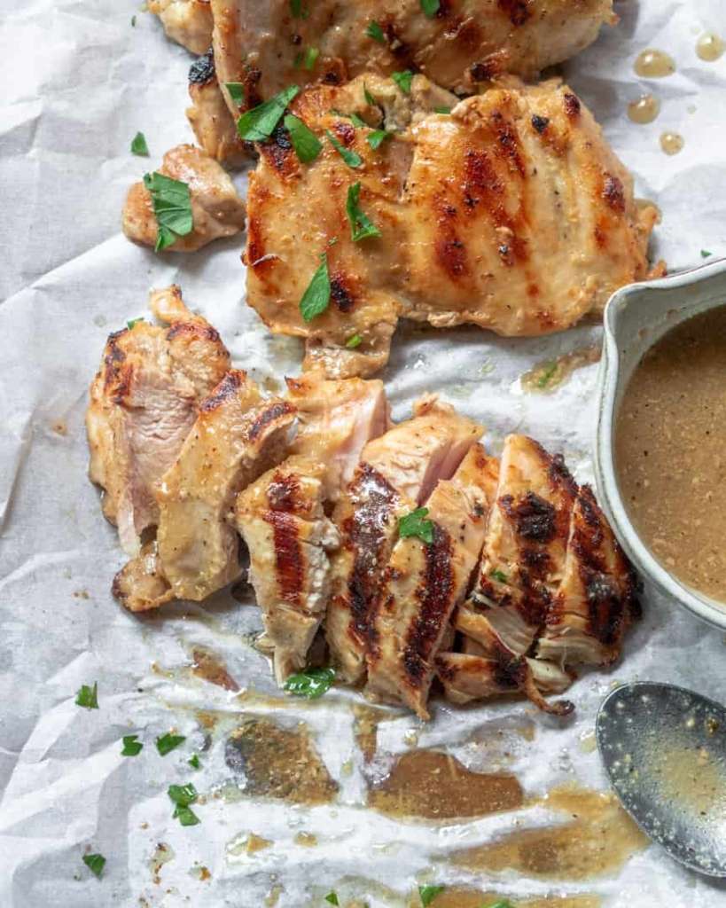 chicken thighs sliced on a baking sheet with a spoon next to it and a small bowl of dressing.