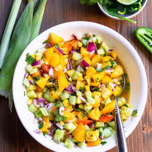 Peach Salsa in a bowl with a spoon in its, surrounded by lime, corn husks, jalepeño.