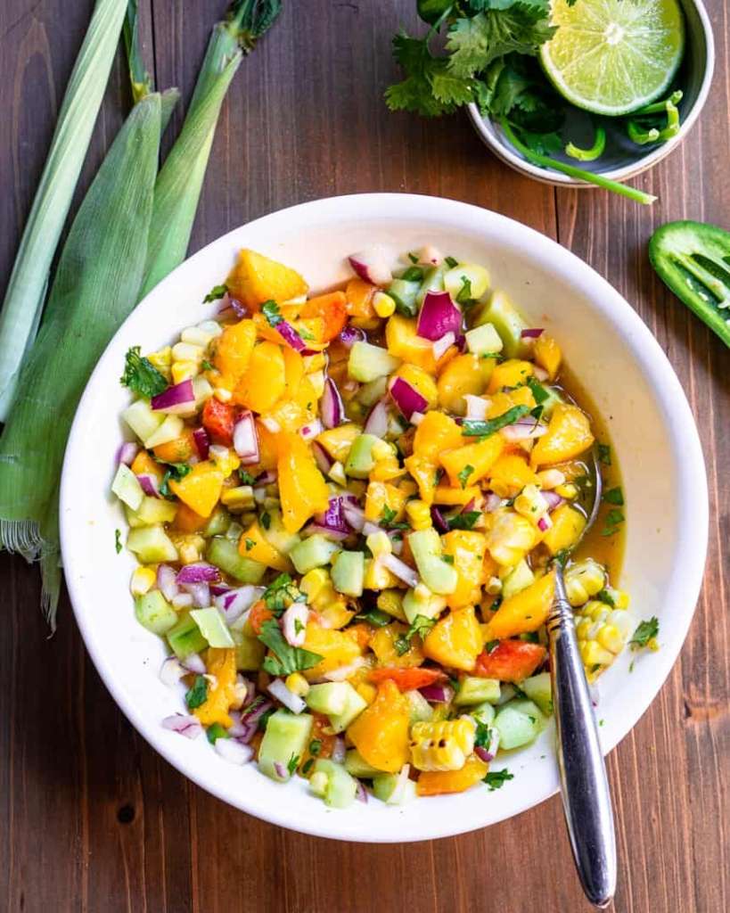 Peach Salsa in a bowl with a spoon in its, surrounded by lime, corn husks, jalepeño.