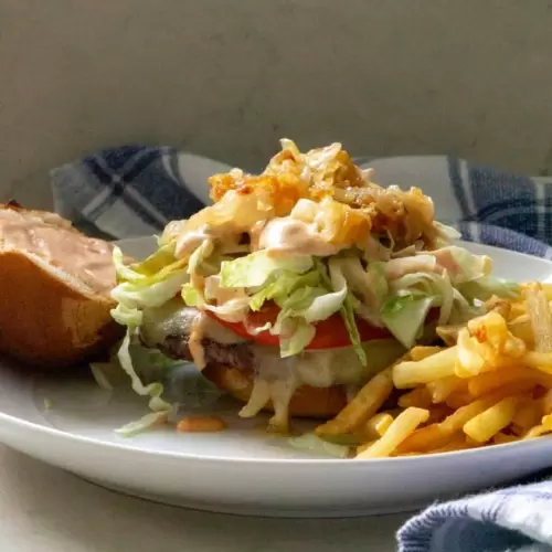 A burger with toppings on a plate with the top of the bun to the side and french fries next to it.