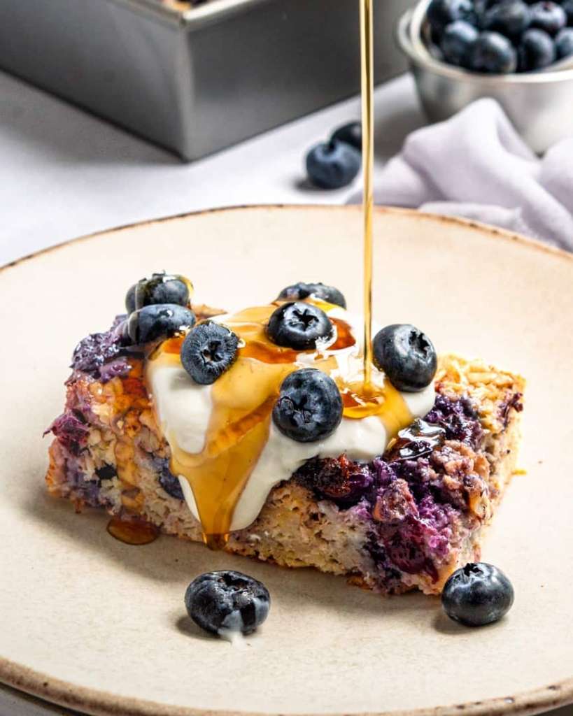 Maple syrup being drizzled over baked oatmeal square with yogurt and fresh blueberries on top.