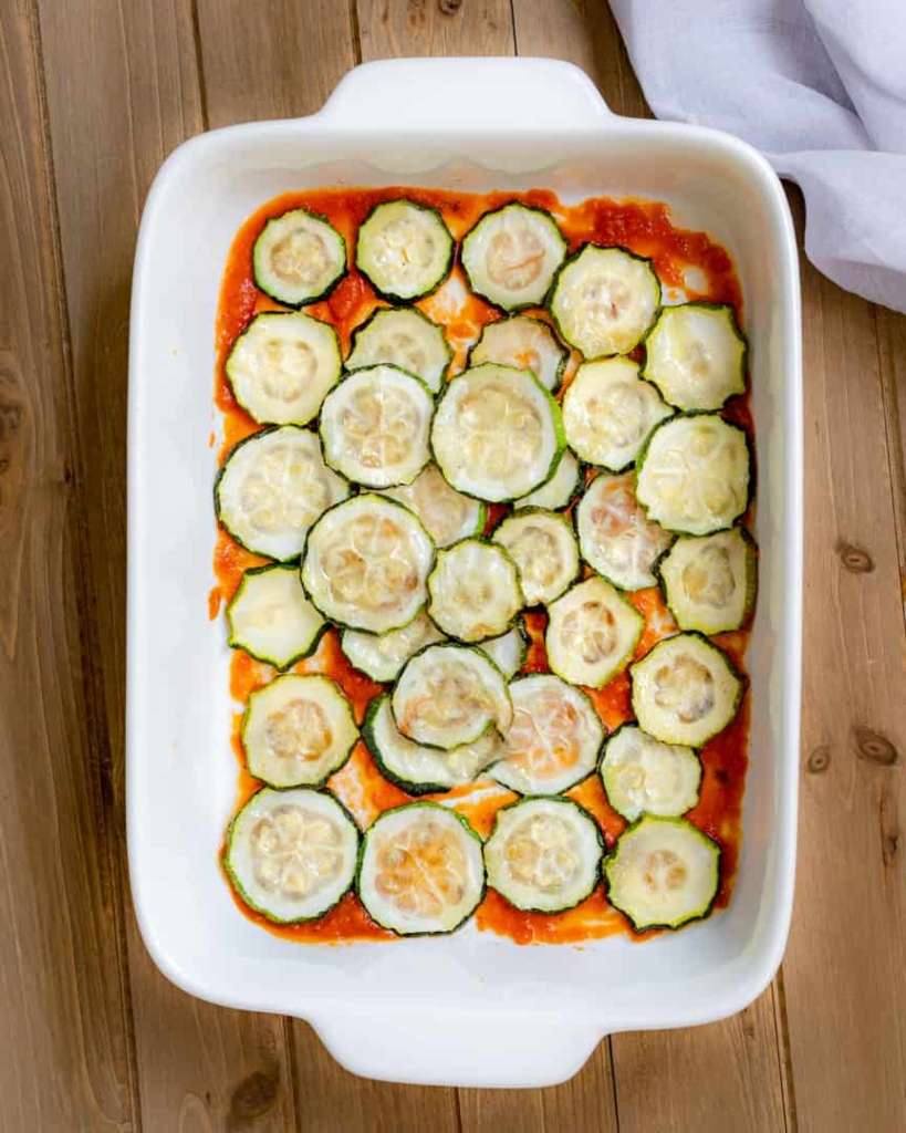 A baking pan with a layer of tomato sauce on the bottom and then a layer of zucchini rounds on top of the sauce.