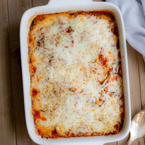 A casserole dish with layered zucchini, sauce and topped with mozzarella cheese. A serving spoon next to the casserole pan.