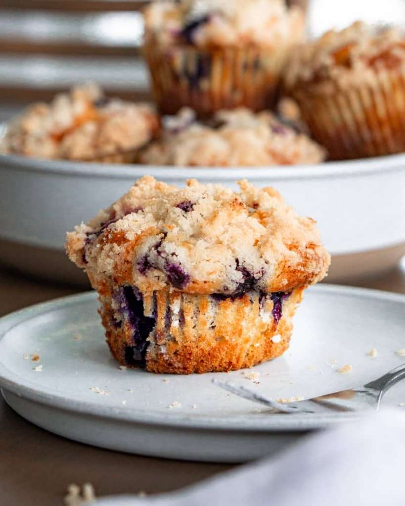 Blueberry muffin on a plate with a fork next to it. A plate of blueberry muffins stacked in the background.