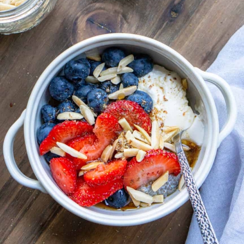 a bowl filled with chia pudding and topped with fresh blueberries, strawberries, yogurt and slivered almonds. The bowl has a spoon in it and a napkin next to it.