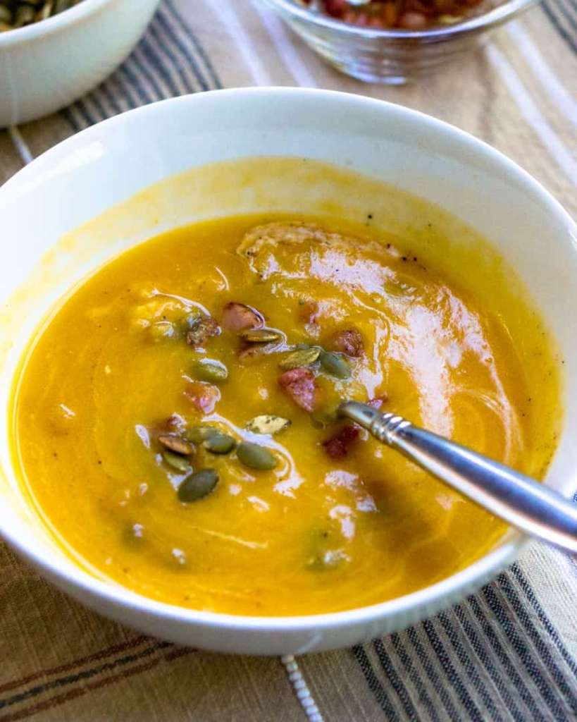 A bowl of butternut squash soup topped with pumpkin seeds and pancetta. There is a spoon in the bowl.