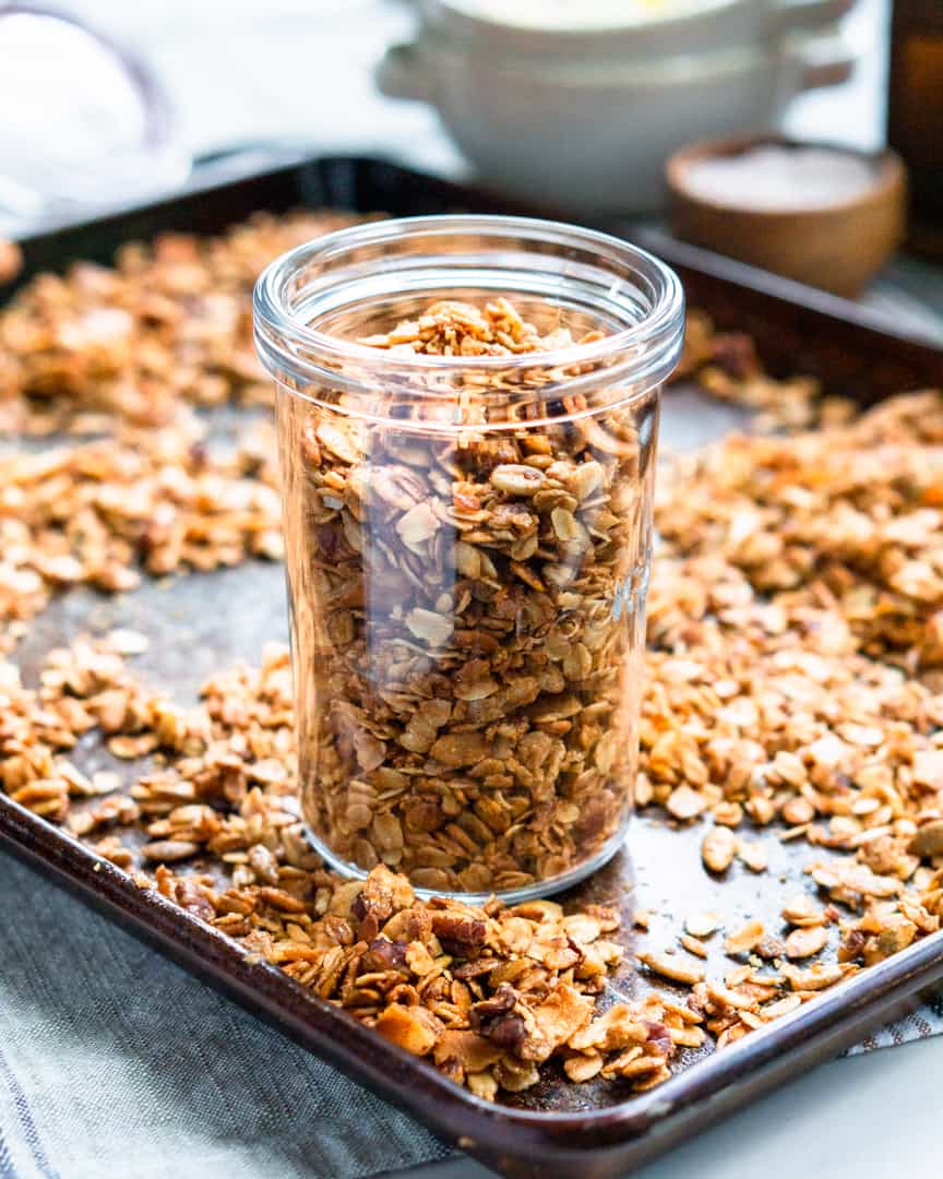 A glass jar filled with granola on top of a baking sheet, surrounded by granola.