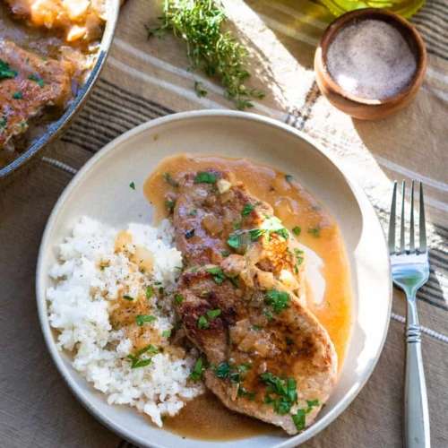 A plate with 2 pork chops with rice next to it. There is pan sauce on the plate and a fork next to it.