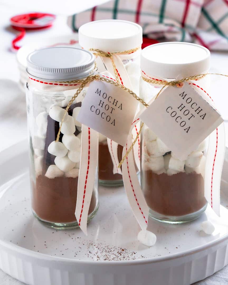 Three tall jars with lids ribbons and tag that are filled with hot cocoa mix and mini marshmallows.