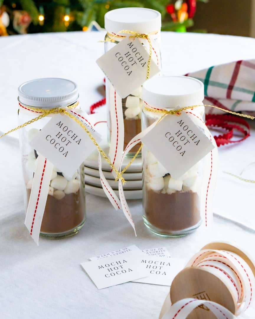 Three jars filled with hot cocoa mix, marshmallows, and chocolate squares. The jars have lids and are embellished with ribbon and a tag with it's name and directions.