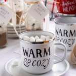A mug on a plate filled with hot cocoa topped with mini marshmallows. The mug says warm and cozy on it and it's surrounded by hot cocoa mix and other mugs.