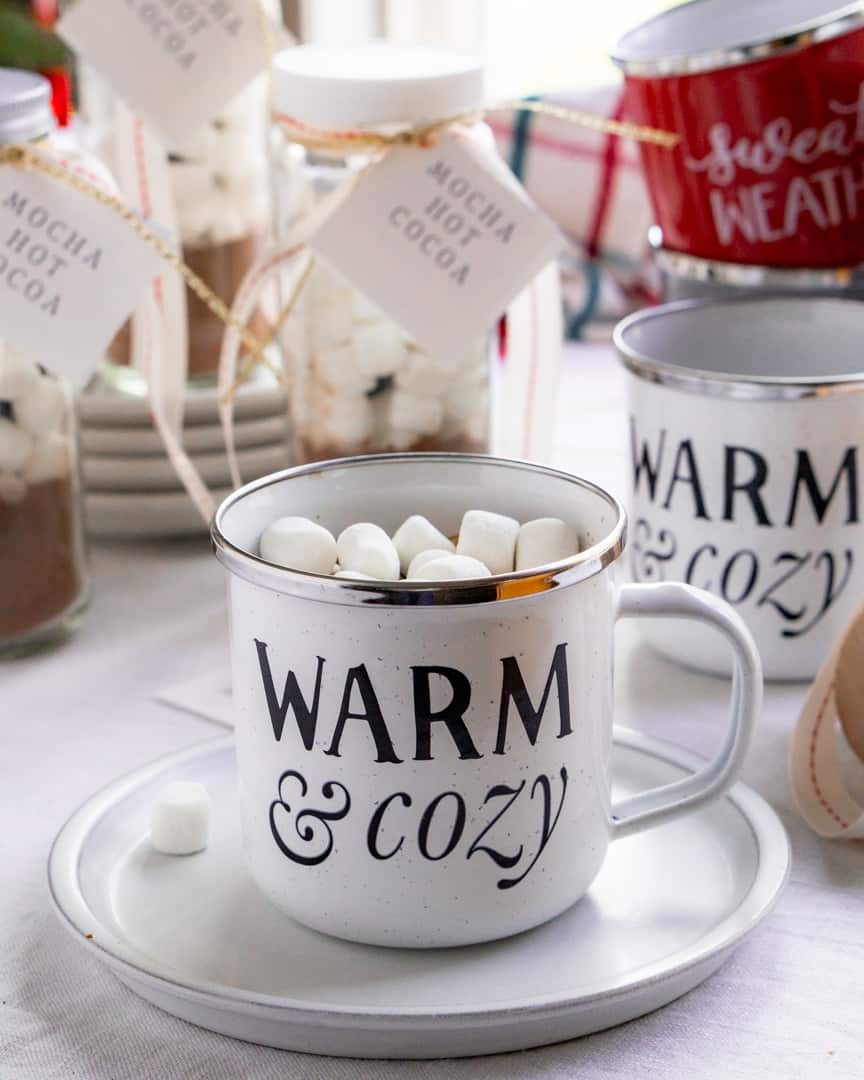 A mug on a plate filled with hot cocoa topped with mini marshmallows. The mug says warm and cozy on it and it's surrounded by hot cocoa mix and other mugs.