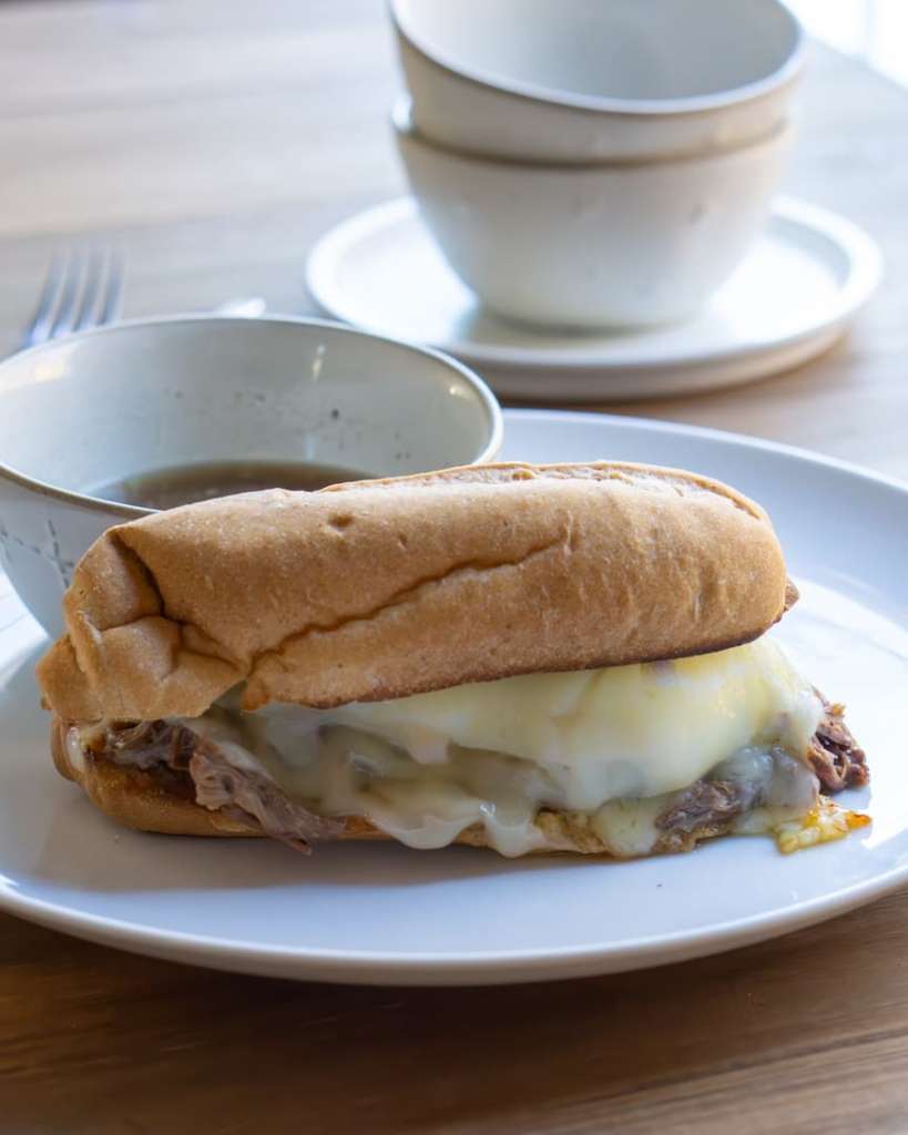 French dip sandwich with melted cheese on a plate with a small bowl of au jus behind it. There are also stacked bowls behind the sandwich.
