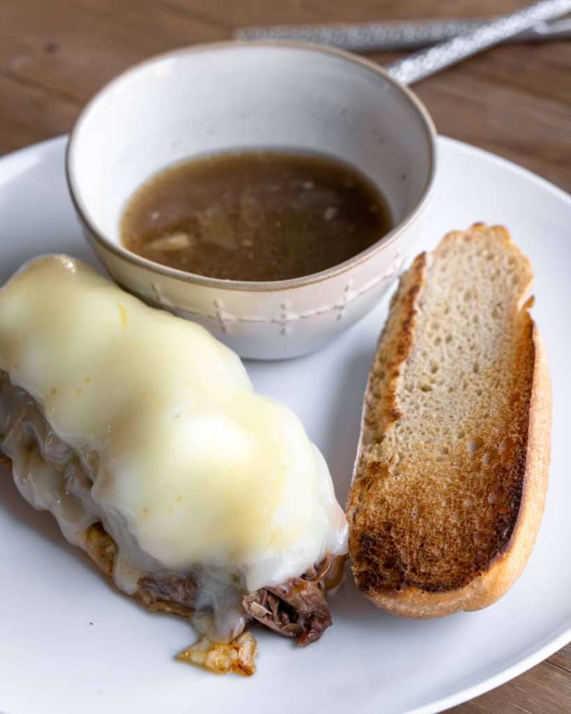 Open faced French dip sandwich with melted cheese and a small bowl of au jus next to it.