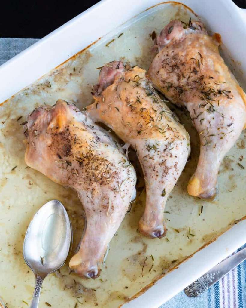 Three baked chicken quarters laying next to each other in a baking pan with a spoon for pan juices.