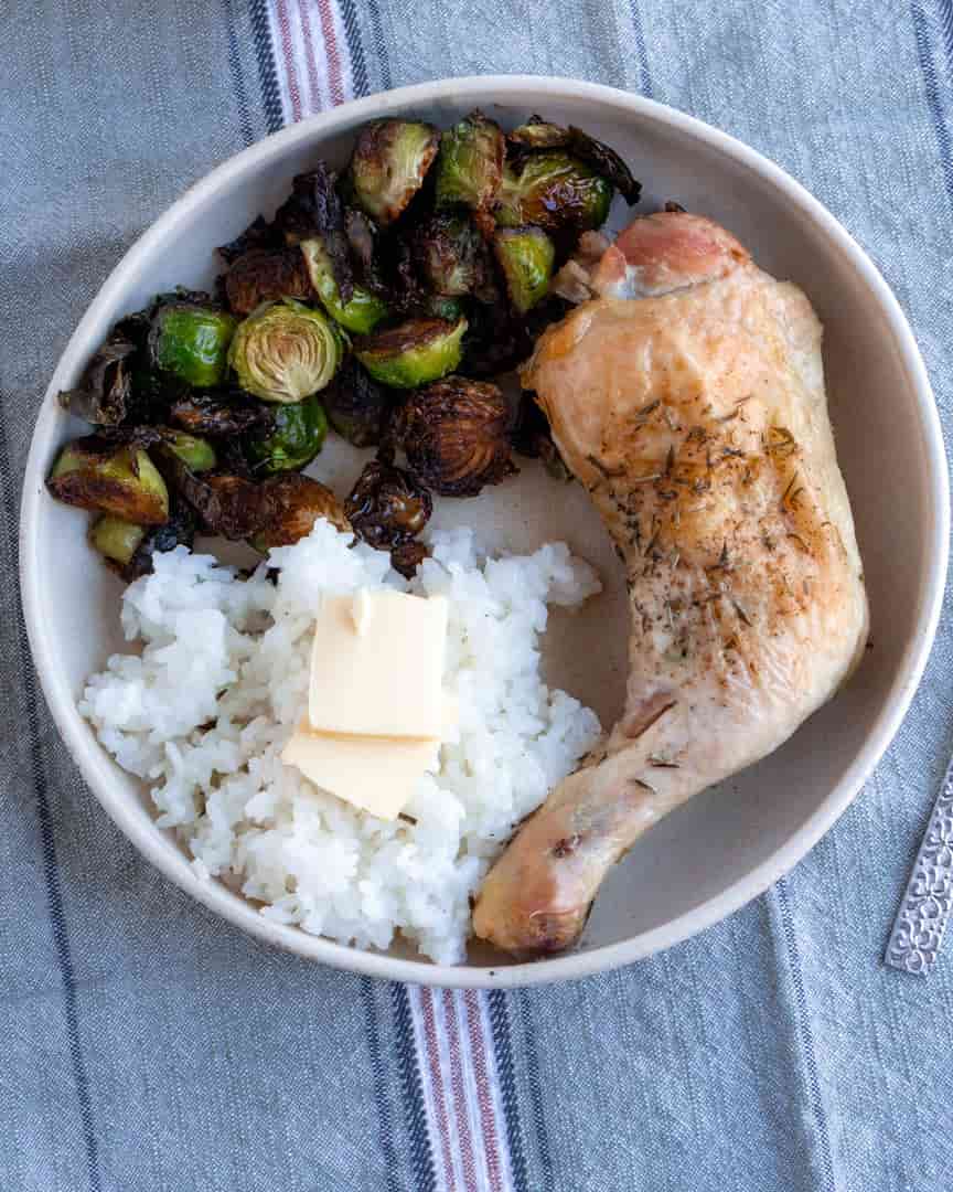 A chicken leg on a plate with rice topped with butter and a side of roasted brussel sprouts
