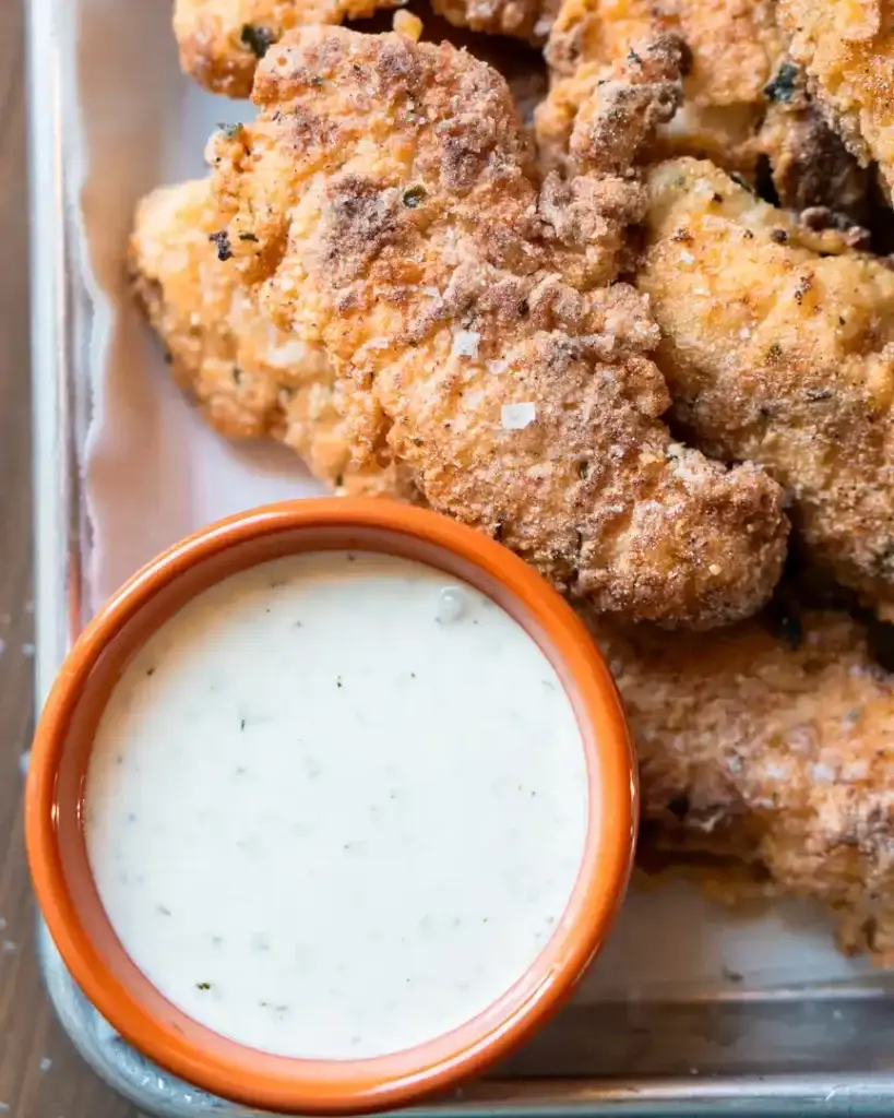 A small bowl of ranch dressing next to fried chicken tenders