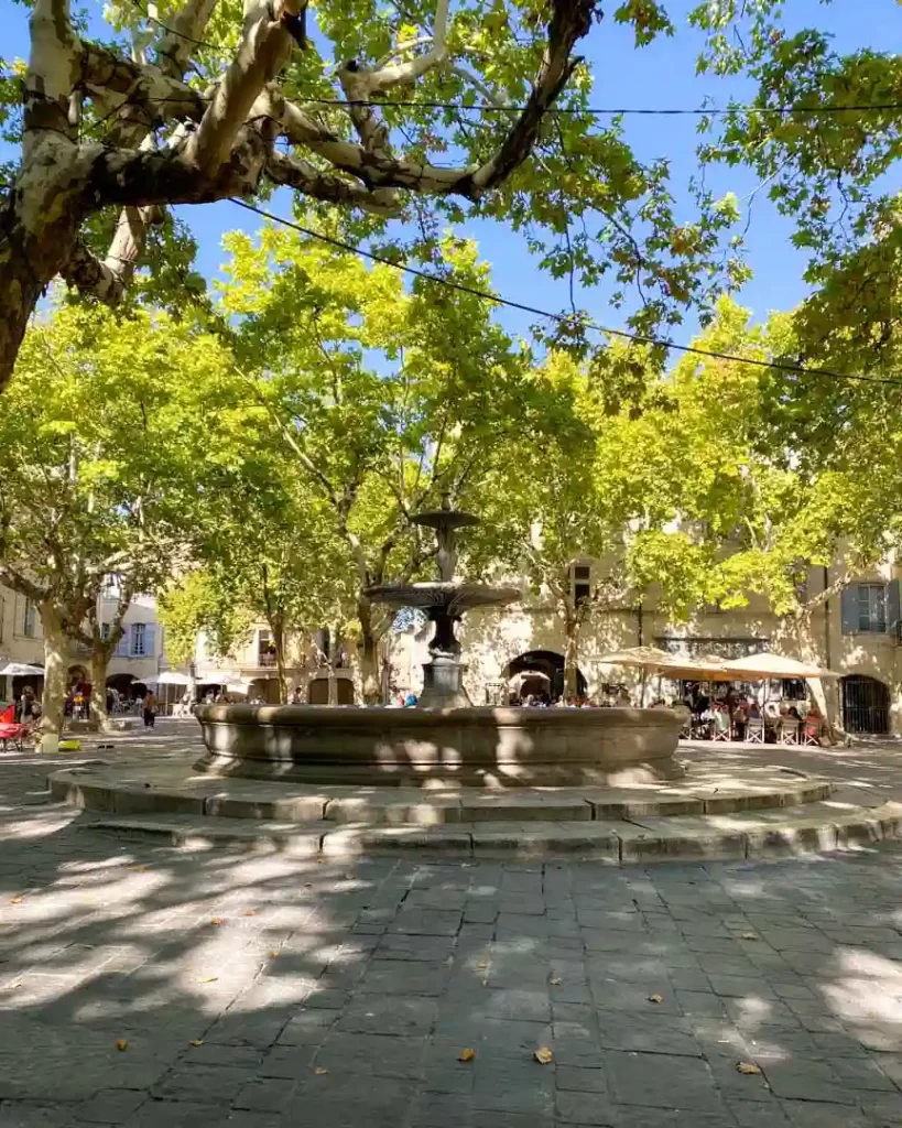 A town square with trees and a large tiered fountain in the middle.