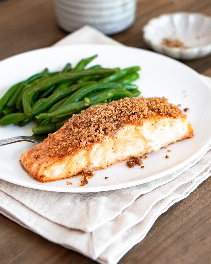 A salmon fillet with brown sugar crust on top it. There are green beans also on the plate with the salmon.