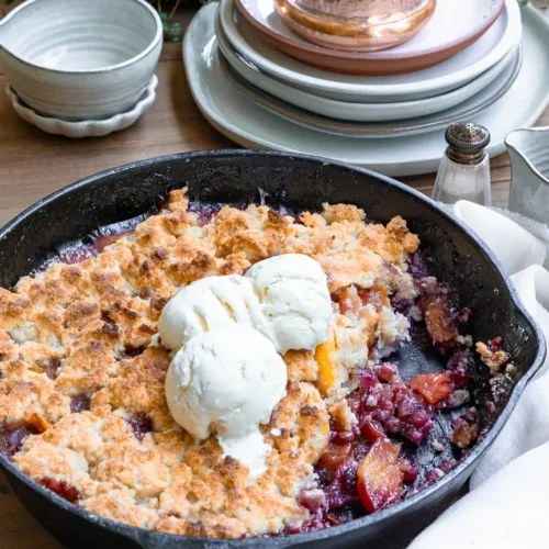 A blueberry peach cobbler with some of it scooped out. There are 3 balls of ice cream on top of the cobbler.