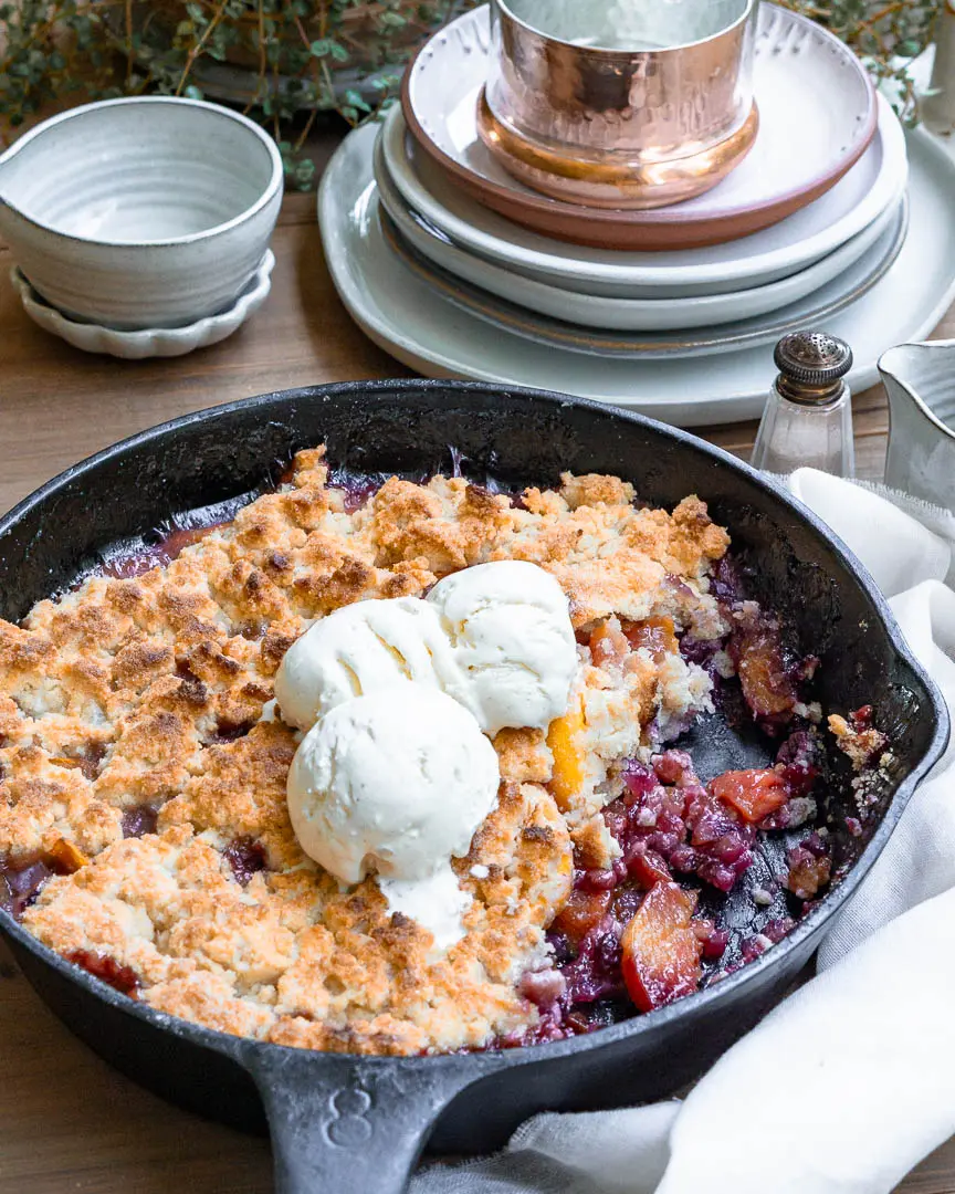 A blueberry peach cobbler with some of it scooped out. There are 3 balls of ice cream on top of the cobbler.