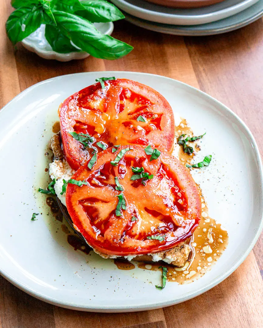 A plate of toast with ricotta cheese and two slices of tomatoes on top of it. There are fresh basil slivers and a balsamic drizzle on top of it.