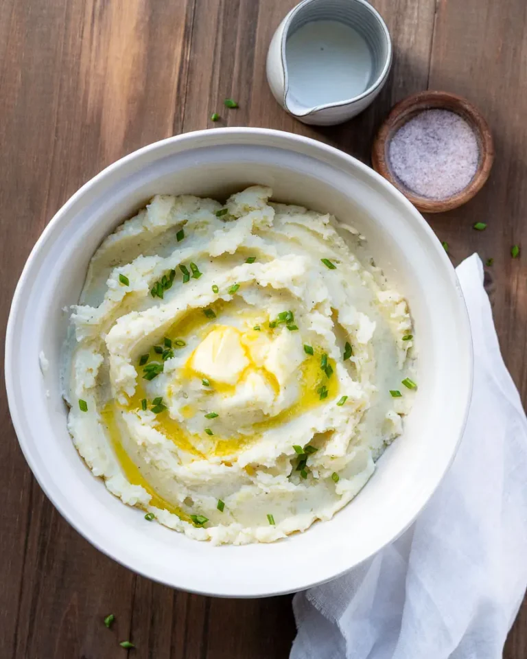 Creamy mashed potatoes in a bowl with slightly melted butter and topped with chives. There is a small pitcher with a bit of cream and a small bowl of salt next to the bowl of mashed potatoes.