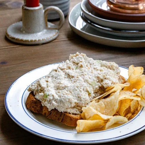 Open faced classic deli style chicken salad sandwich on a plate. There is a side of potato chips on the plate.