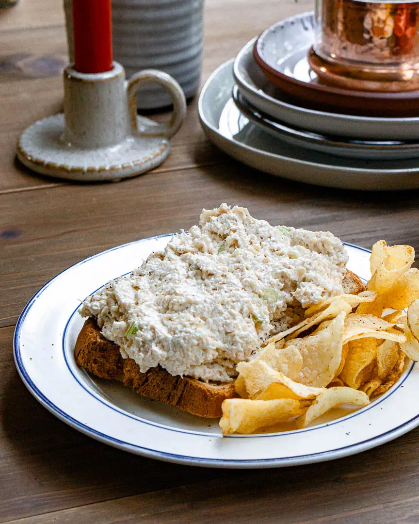 Open faced classic deli style chicken salad sandwich on a plate. There is a side of potato chips on the plate.