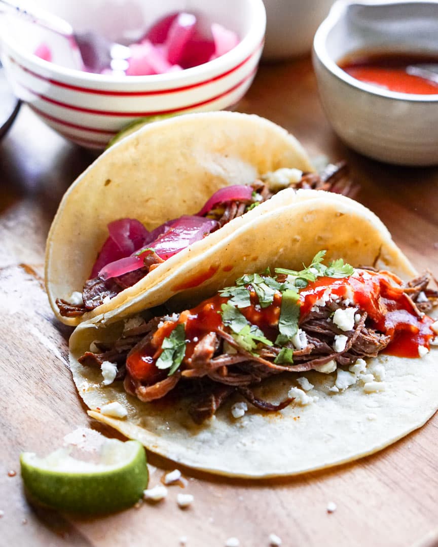 Two brisket tacos propped next to each other. There is a squeezed wedge of lime next to them.