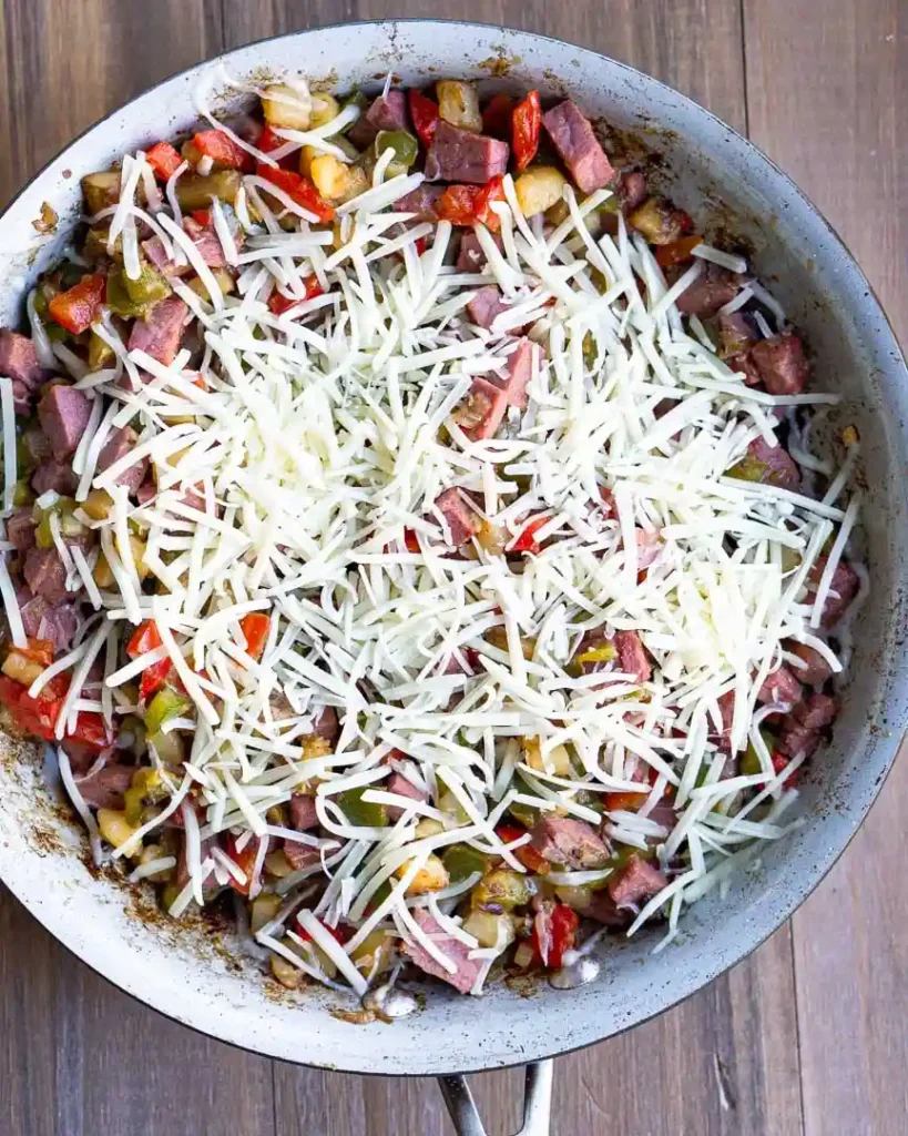 Shredded cheddar cheese on top of corned beef hash in a skillet.