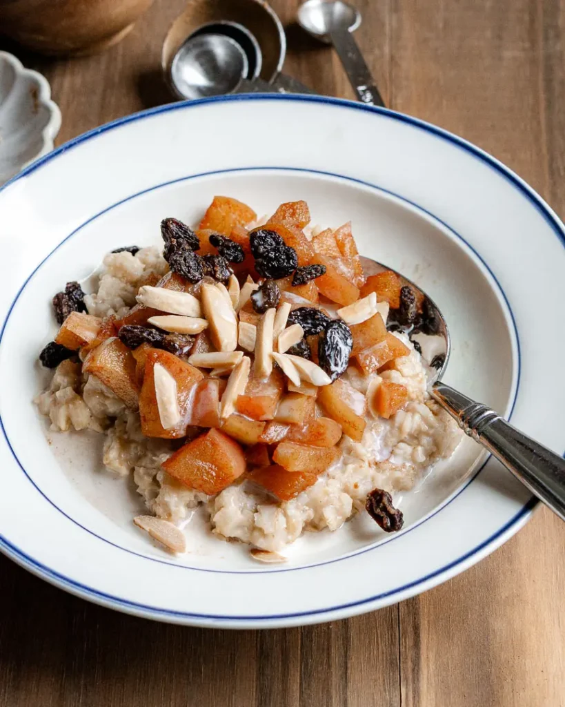 A bowl of apple cinnamon oatmeal with slivered almonds and raisins on top. There is also a spoon on the bowl.