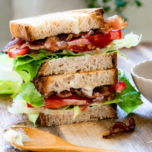 Blt sandwich cut in half and each half is stacked on top of the other. There is a small wooden spoon with some garlic mayo on it that sits in front of the sandwich.