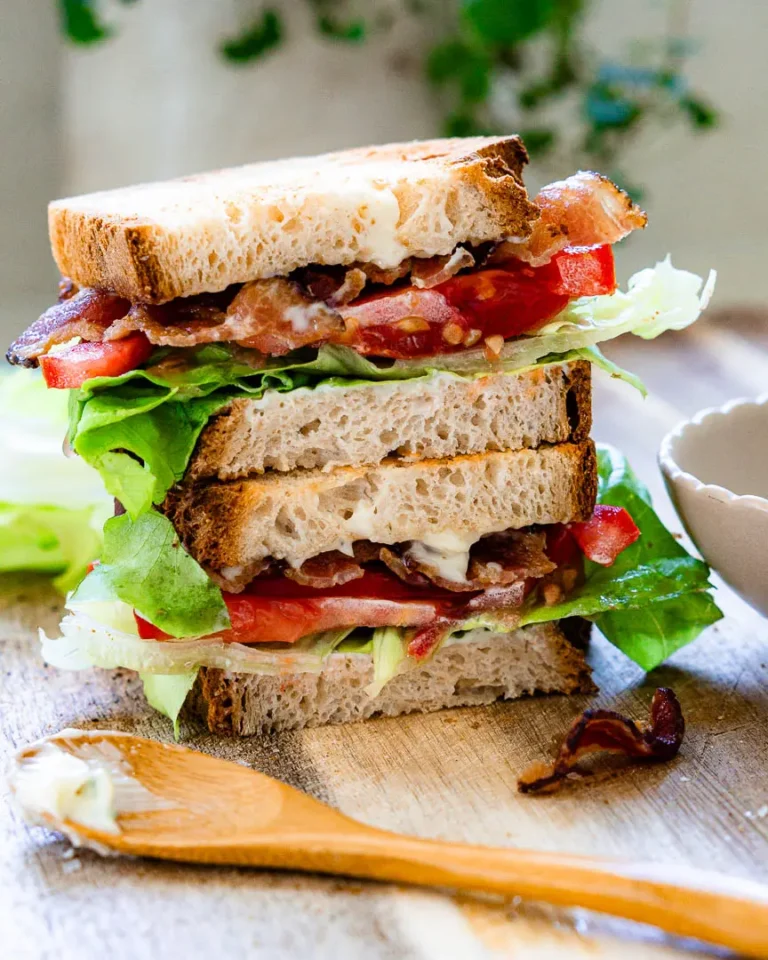 Blt sandwich cut in half and each half is stacked on top of the other. There is a small wooden spoon with some garlic mayo on it that sits in front of the sandwich.