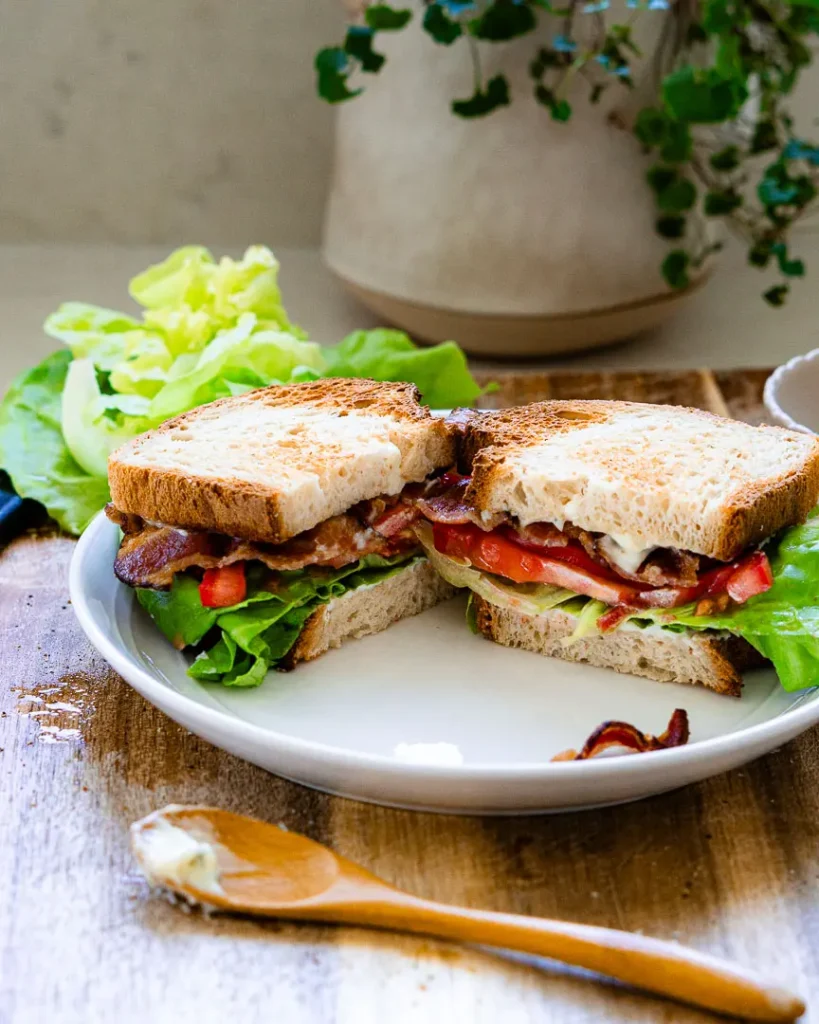 A blt sandwich cut in half on a plate. There is a small spoon with garlic mayo sitting in front of the plate.