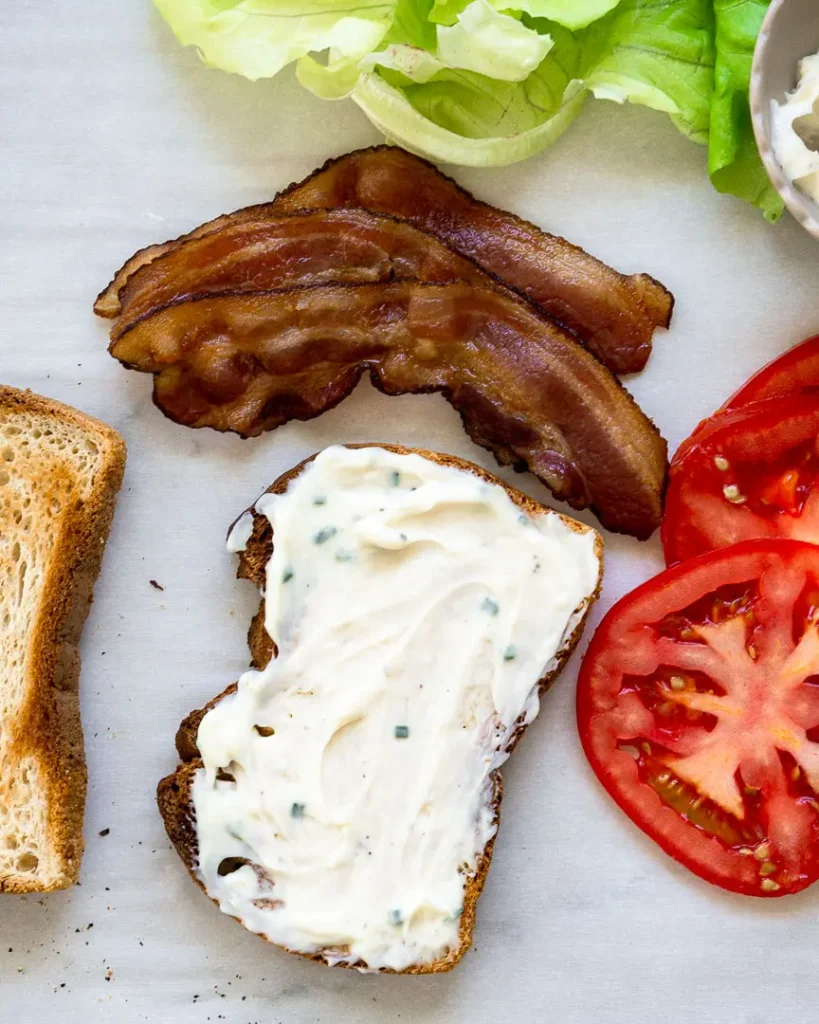 One slice of bread coated with garlic mayo. There are sliced tomatoes, lettuce, and bacon next to the slice of bread.