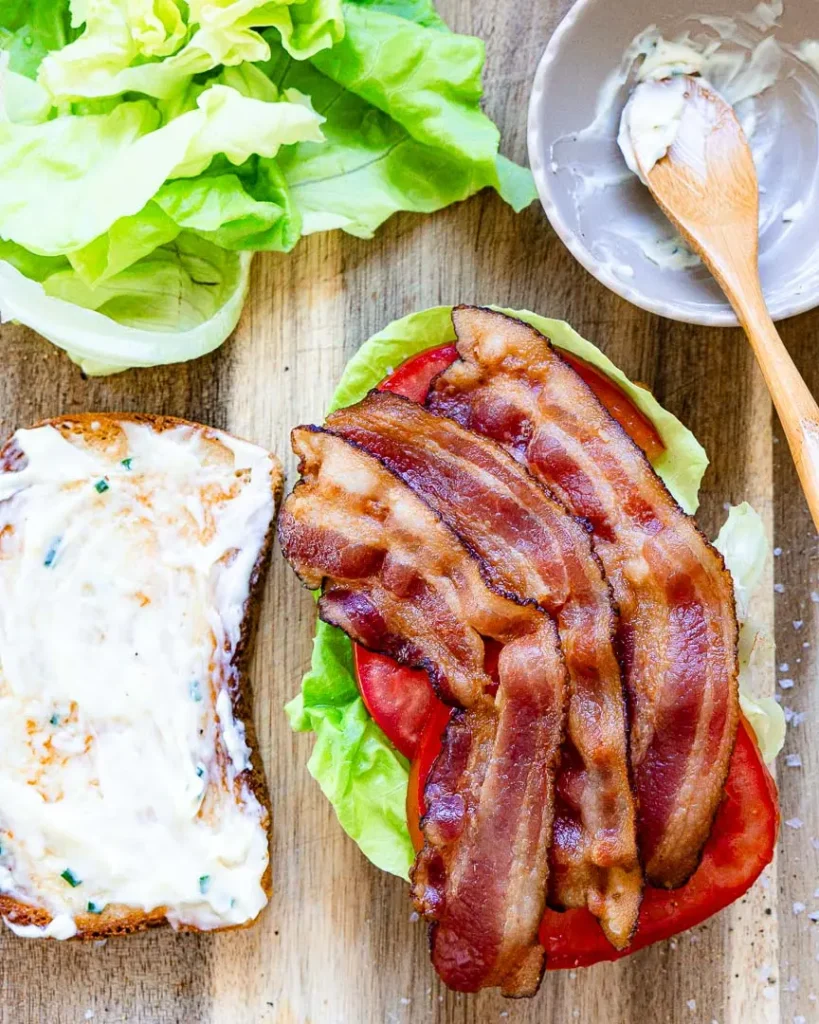 Bacon sitting on top of half of a blt sandwich. There is a slice of bread with garlic mayo next to it.
