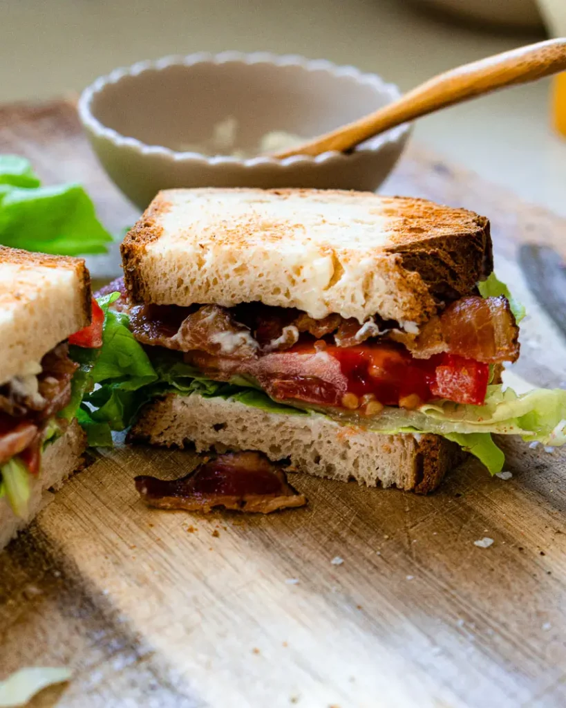 A cut in half blt sandwich where you can see the inside of the sandwich. There is a small bowl behind the sandwich with a small wooden spoon sitting in it.