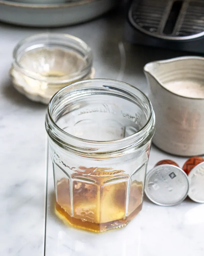 An empty jam jar with maple syrup and brown sugar at the bottom of it. There is a small pitcher of milk and 2 Nespresso pods next to the jar.