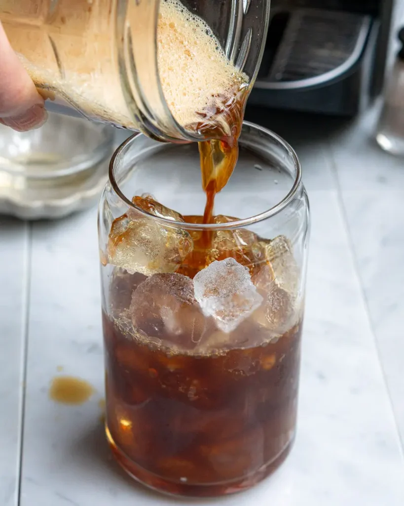 Sweetened espresso being poured over a tall glass filled with ice.