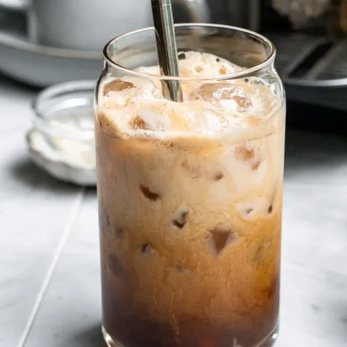 A tall glass of iced French toast latte with a metal straw sticking out of it. there is a Nespresso machine behind the glass.